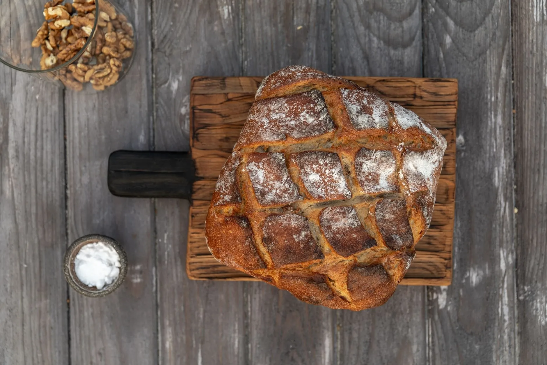 A scored country loaf on a wooden board with a small dish of white salt beside it