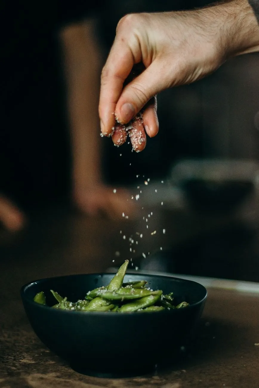A hand sprinkling salt over a bowl of charred edamame