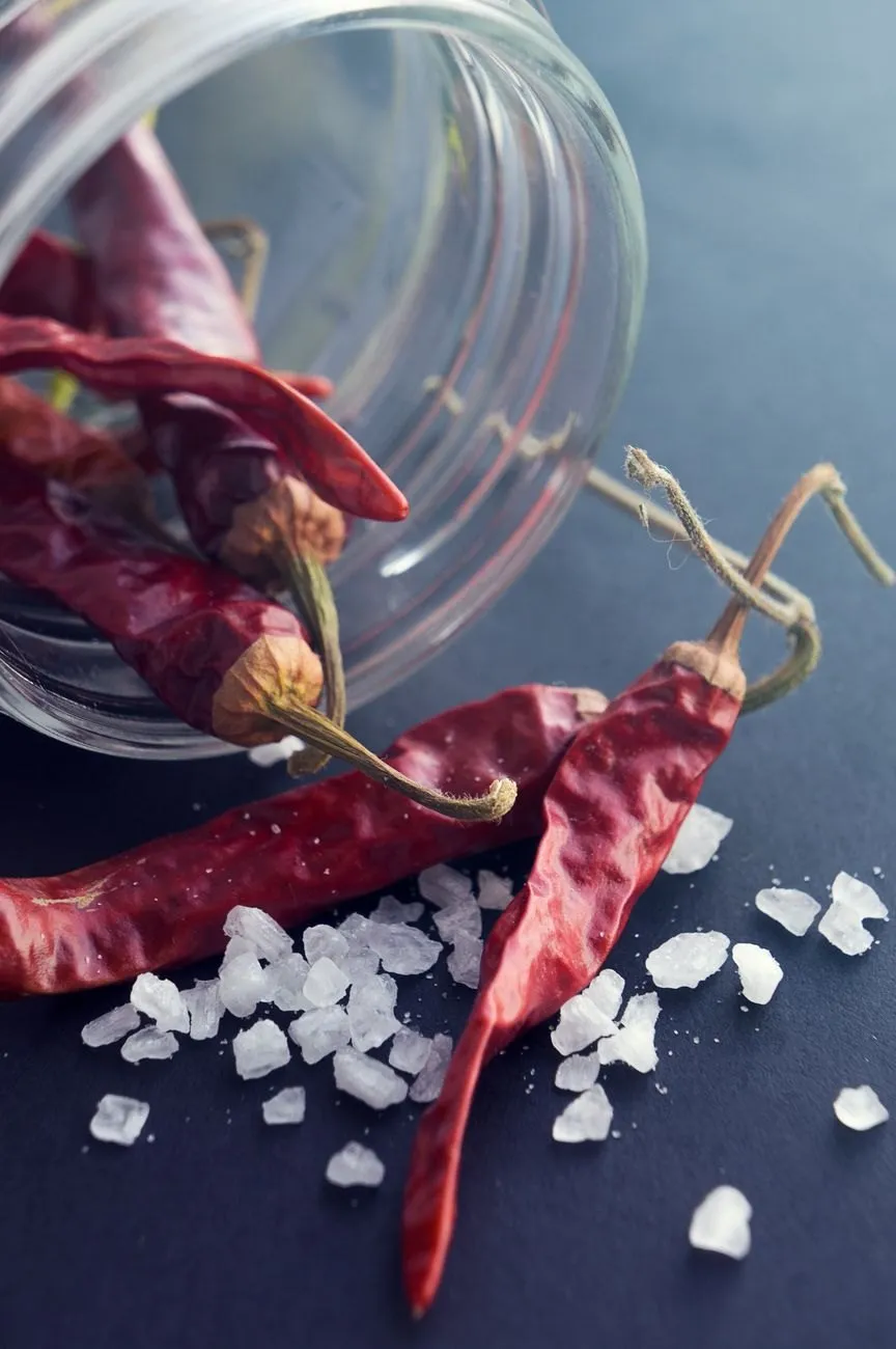 Dried red chillies spilling from a glass jar beside clear crystals of coarse salt