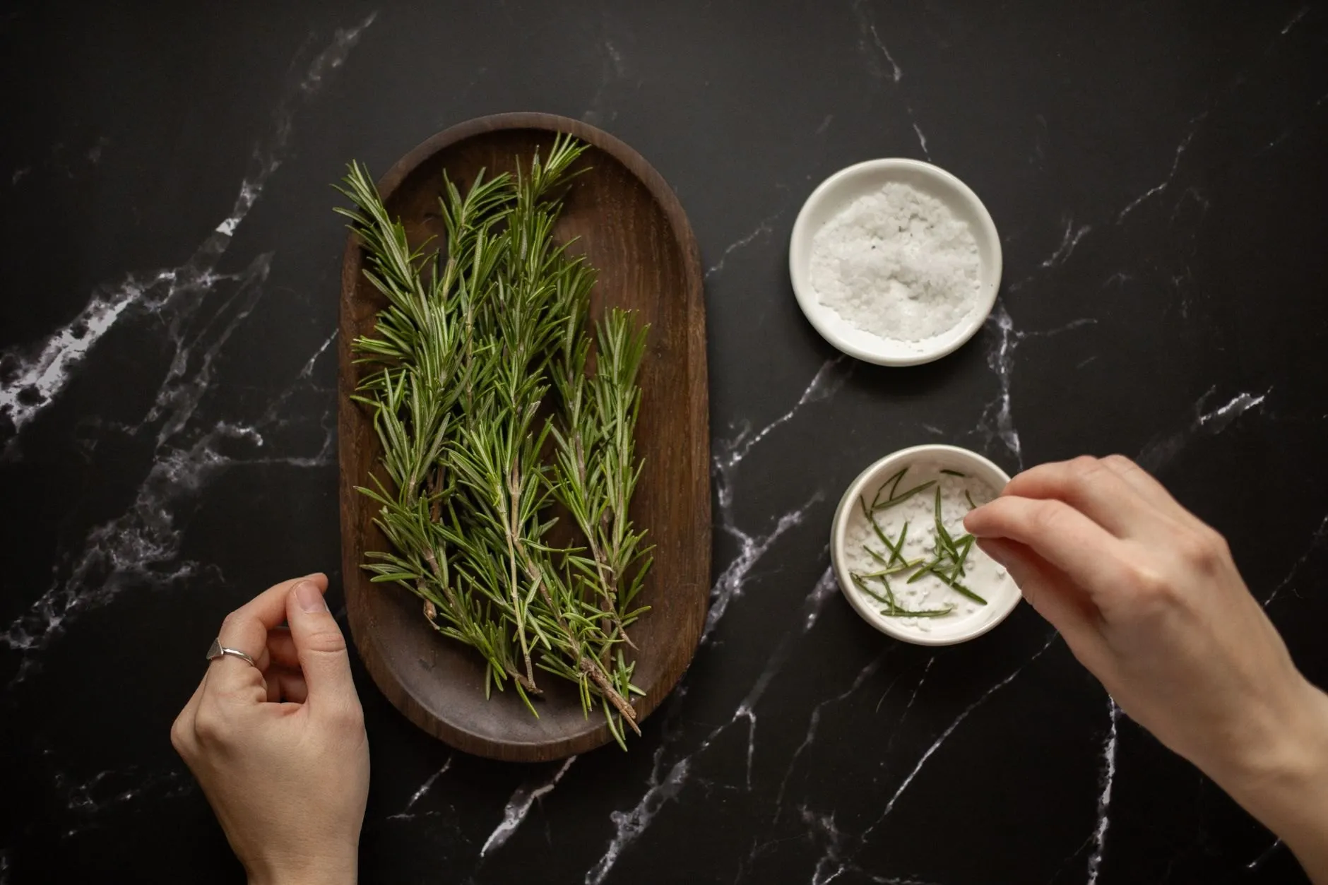 Hands pinching rosemary into a dish of salt beside a wooden tray of fresh rosemary sprigs
