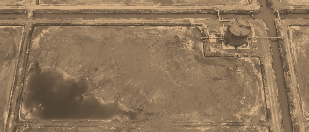 A drained crystalliser from 300 metres holding a shallow film of leftover liquor in its low corner, a drain channel running to a squat storage tank on the bank.