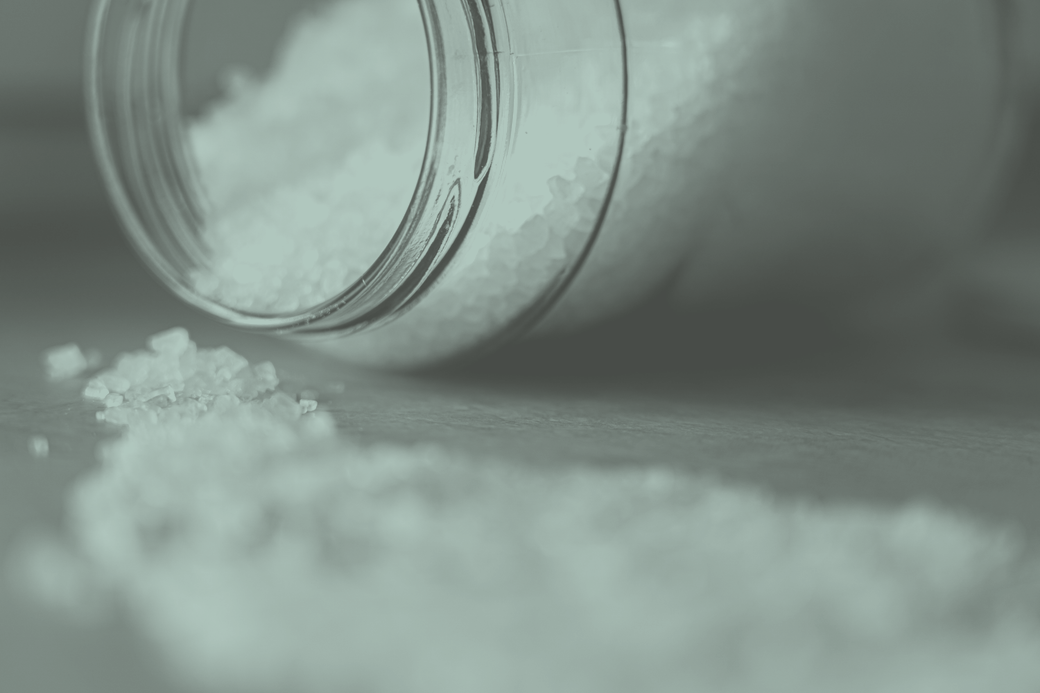 A glass kilo jar tipped on its side with coarse salt spilling out of it onto a plain surface.