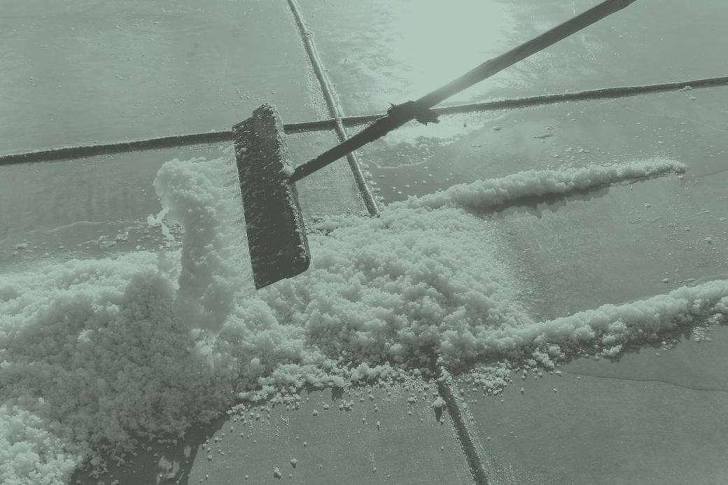 A wooden-bladed rake pushing a ridge of wet salt crystals across the flat floor of a crystalliser pan, with shallow brine standing beyond it.